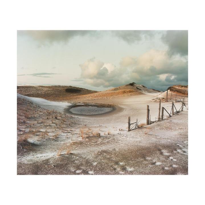 A frozen pond sits in a snowy, hilly landscape with a broken fence under a cloudy sky.
