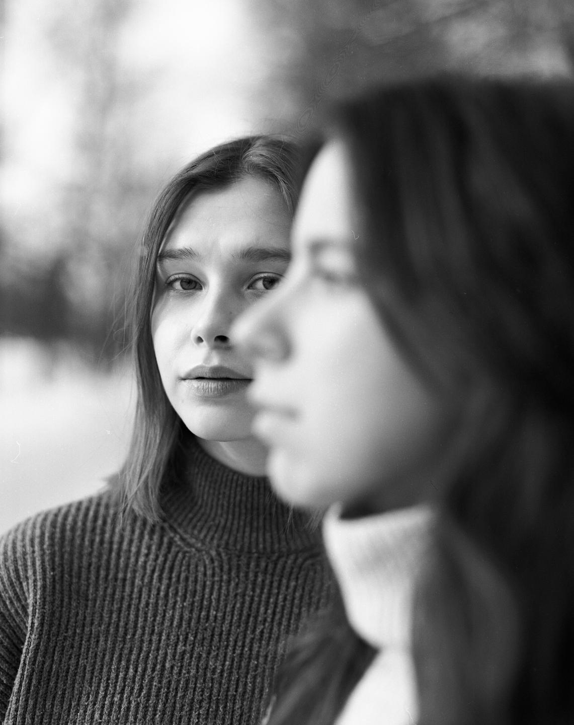 A woman in a turtleneck sweater looks at the viewer, with another woman out of focus in the foreground.