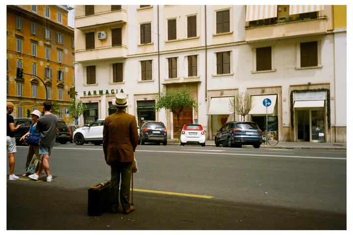 A man in a hat and brown jacket stands with a cane and suitcase on a city street with cars and buildings.