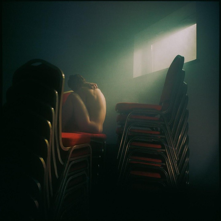 A nude woman sits on a red chair in a dark room with stacks of chairs and a bright window.