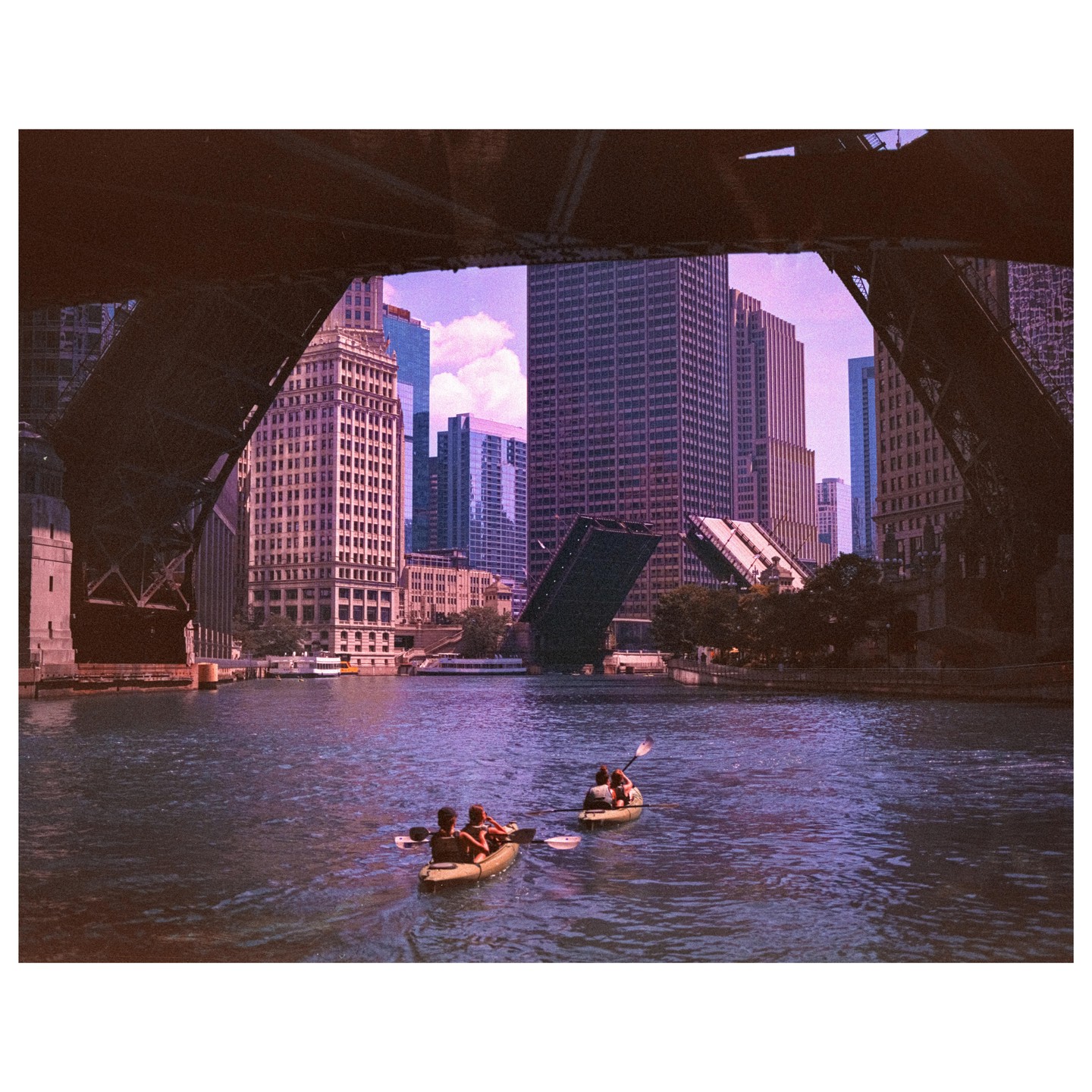 Two kayaks with people paddle on a river framed by a bridge, with city buildings and an open drawbridge in the background.