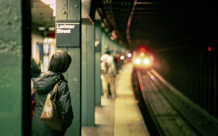 A woman stands on a subway platform at Lorimer Street as a train approaches in the distance.