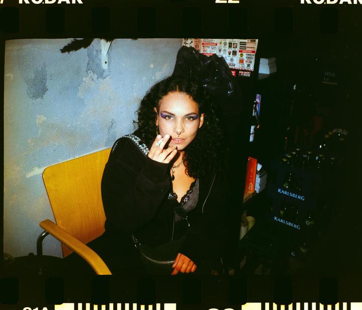 A woman with curly hair and purple eyeshadow sits in a yellow chair, holding a cigarette.