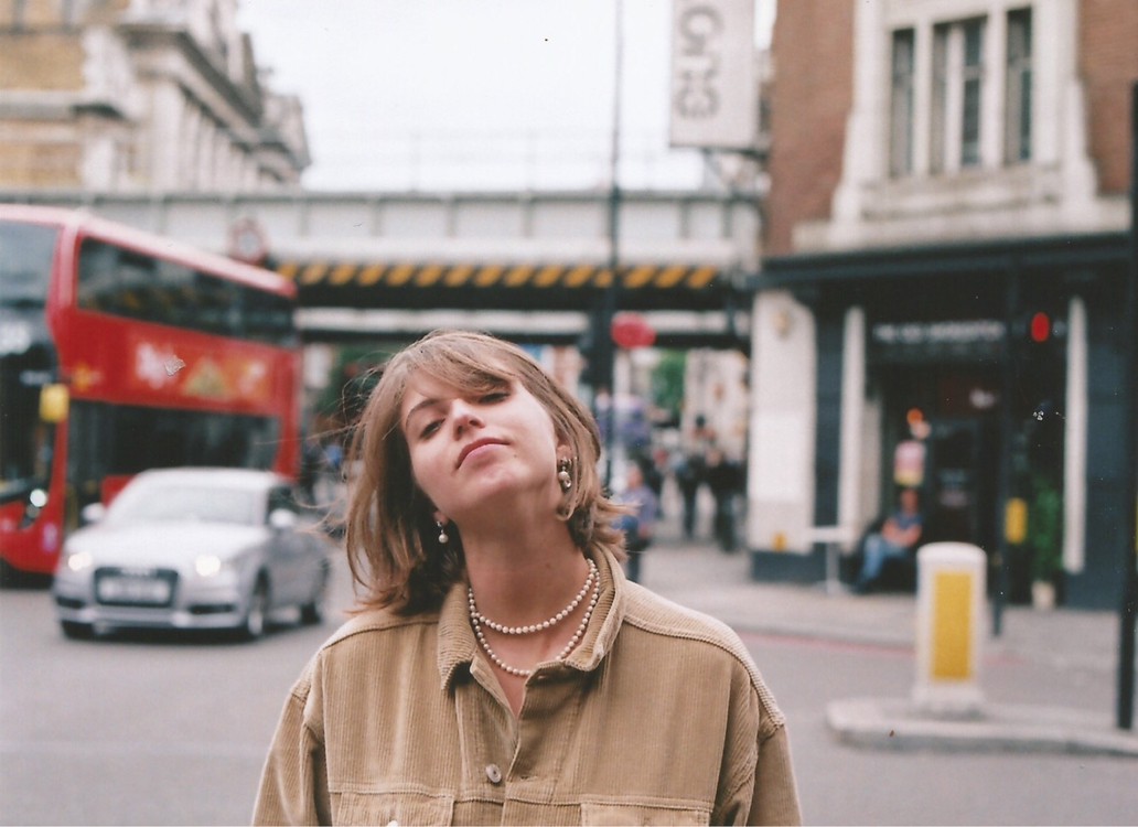 A woman wearing a corduroy jacket and pearl necklace looks at the camera on a city street with a red double-decker bus and cars in the background.