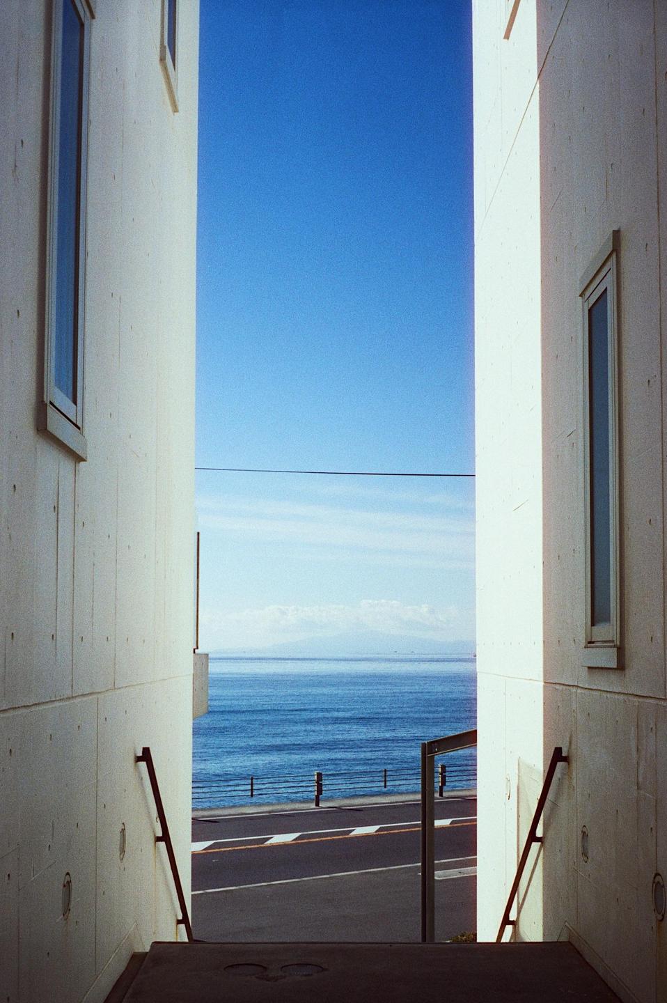 A narrow view between two buildings reveals a road, the ocean, and a distant mountain range under a clear blue sky.