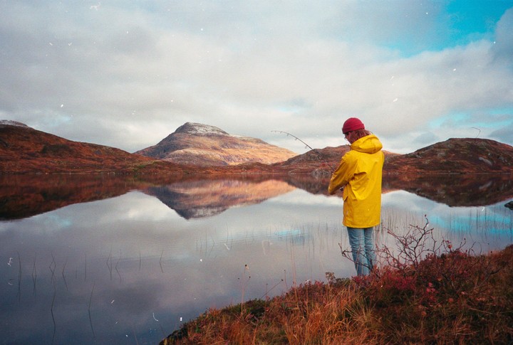 A person in a yellow raincoat and red hat fishes in a lake surrounded by mountains with autumn foliage.