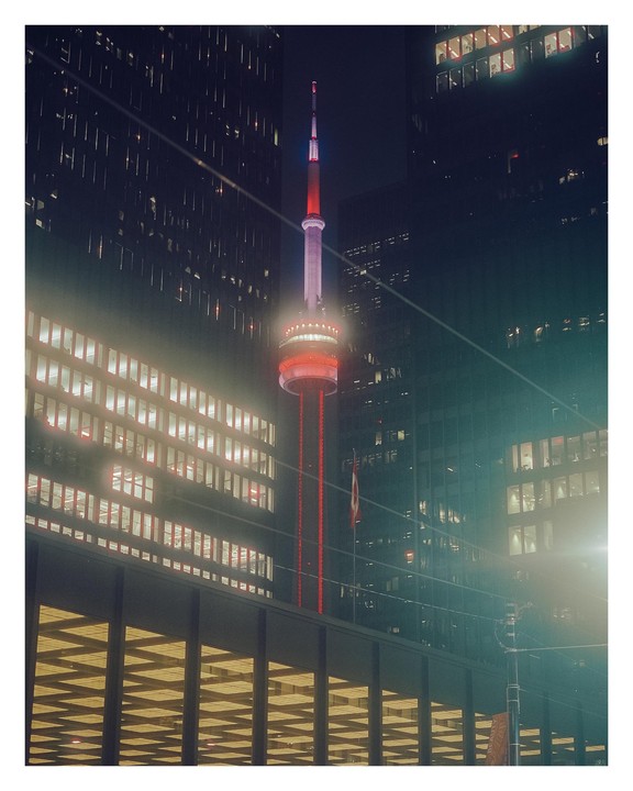 The CN Tower is illuminated in red and white at night, surrounded by tall buildings with glowing windows.