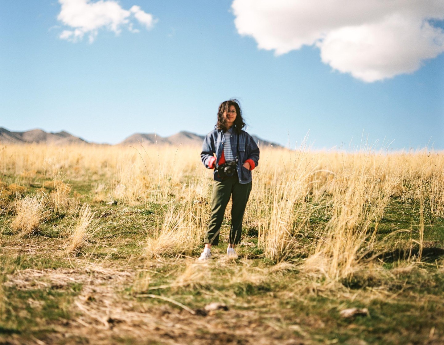 A woman stands in a field of tall, dry grass with mountains in the background under a blue sky with a single cloud.