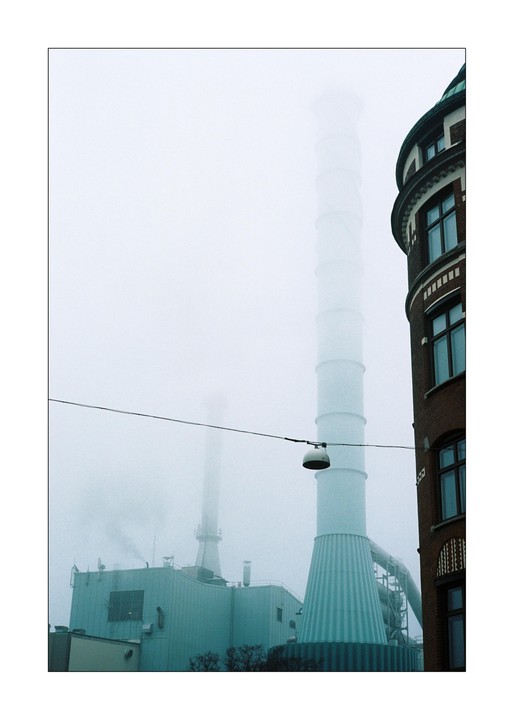 A tall white chimney rises into a misty sky next to a brick building with a rounded corner.