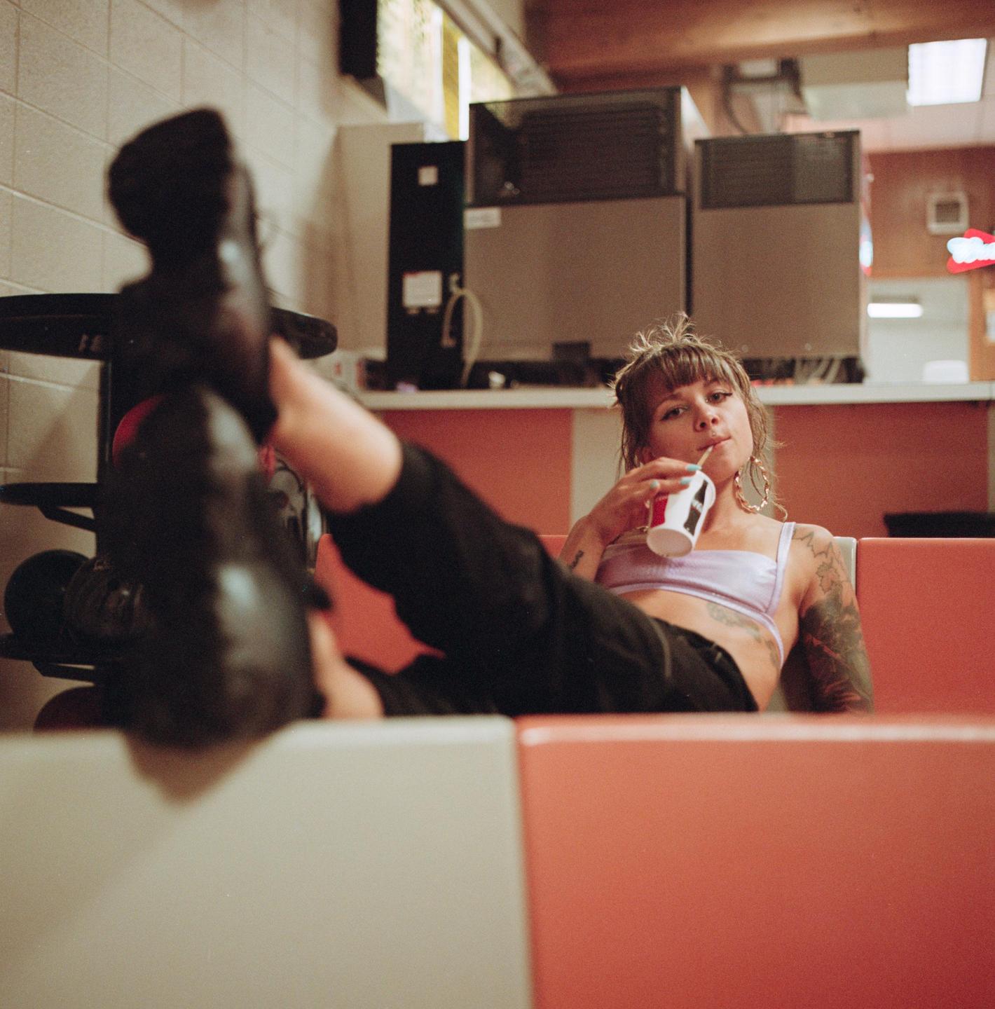 A woman with tattoos on her arms is lying on a red bench with her feet up, drinking from a cup with a straw.