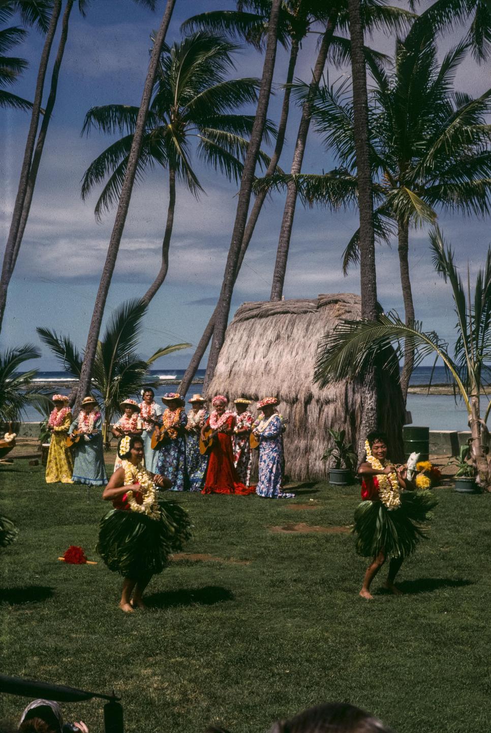 Two women dance in grass skirts and leis while other women in leis and dresses stand behind them near a thatched hut and palm trees.