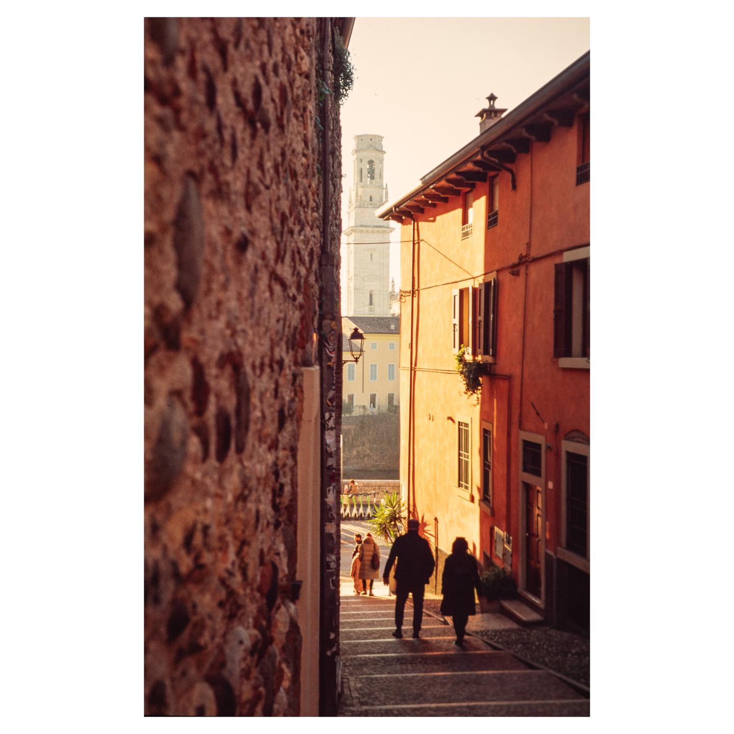 Two people walk down a narrow street with tall buildings on either side, leading to a distant bell tower.