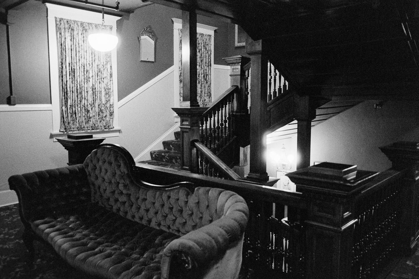 A dark, ornate chaise lounge sits in the foreground of a grand staircase in a dimly lit interior.