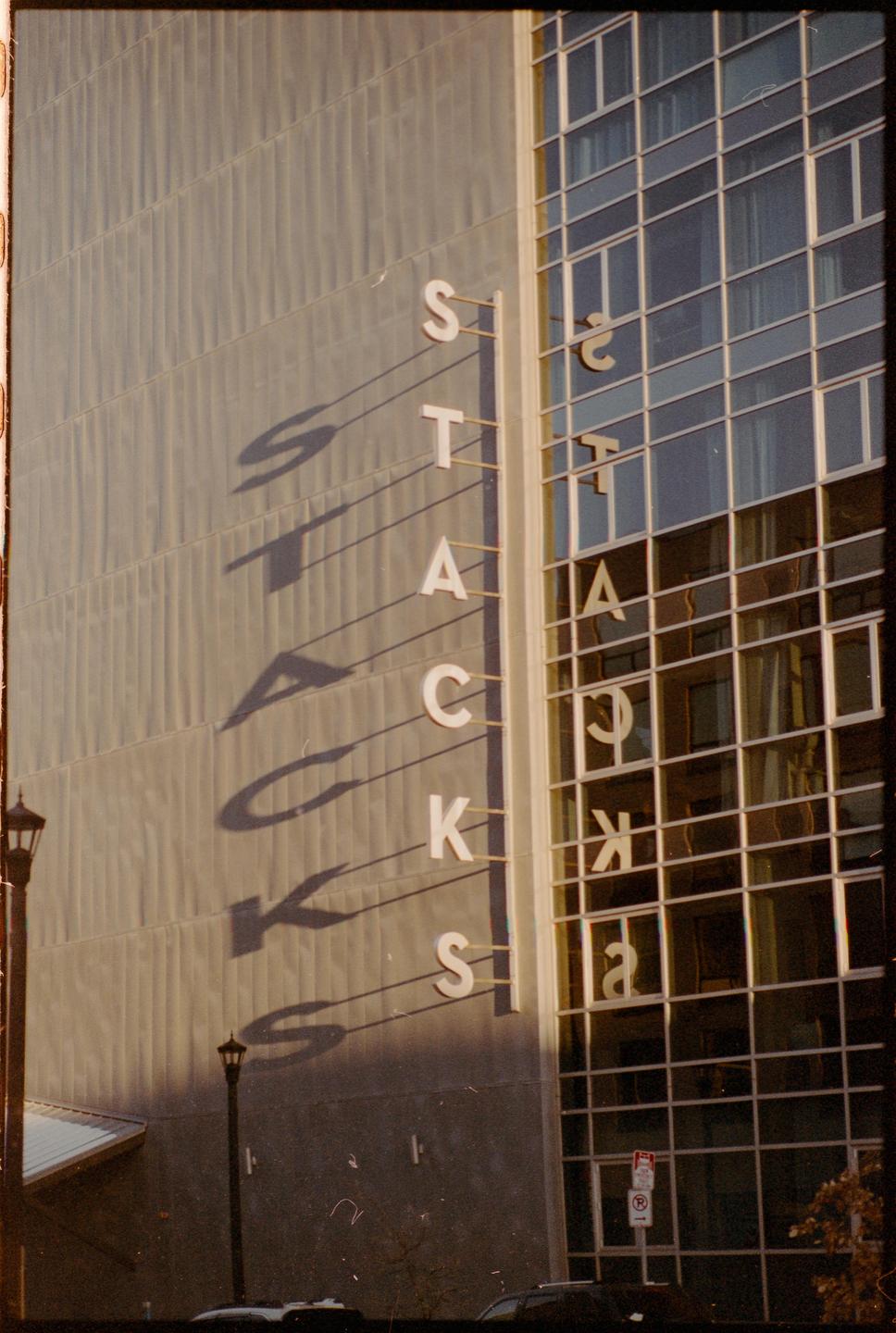 The word "STACKS" is written vertically on the side of a building, casting long shadows on the wall, with its reflection visible in the adjacent windows.