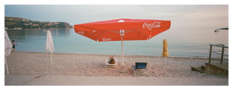 A red Coca-Cola umbrella shades a person relaxing on a beach chair next to the sea.