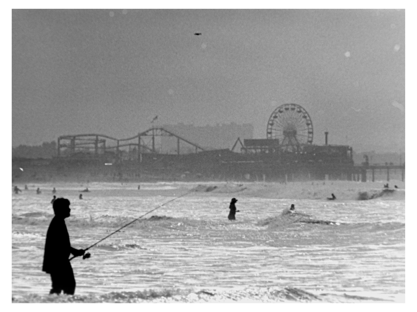 A man fishes in the ocean with a pier and amusement park rides in the background.