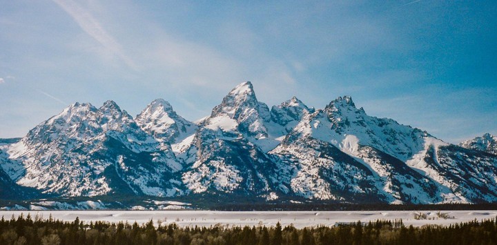 A mountain range with snow-capped peaks rises above a forest and a frozen lake under a clear sky.