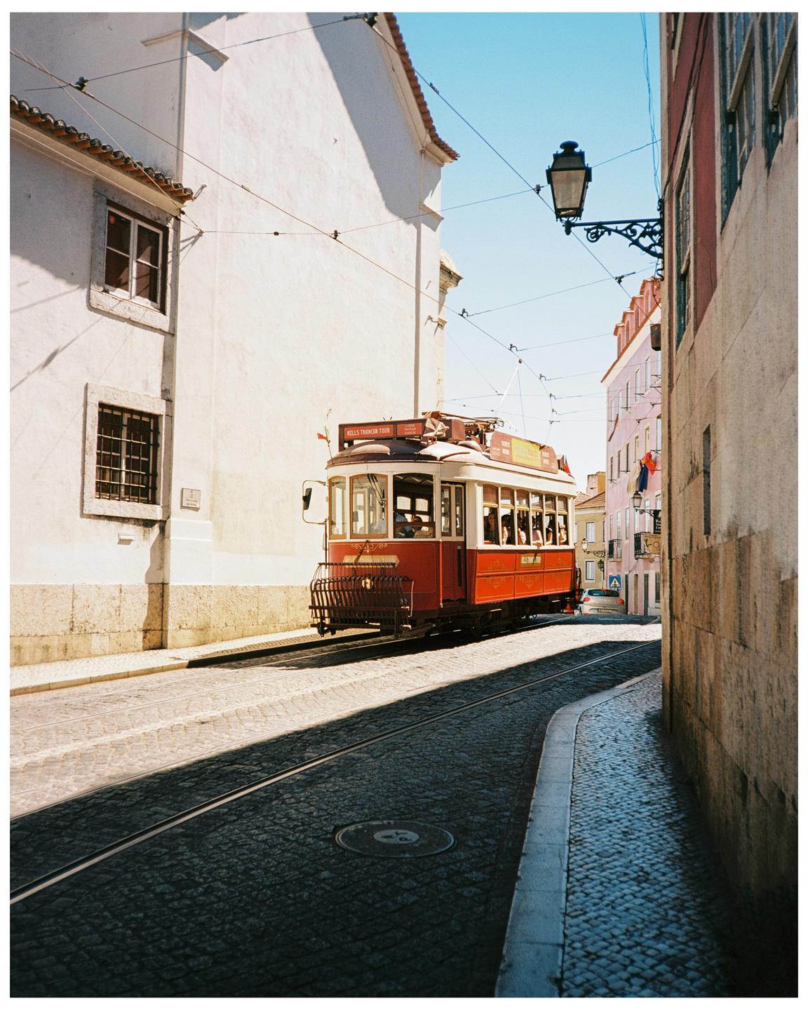 A red tram travels down a narrow cobblestone street lined with buildings.