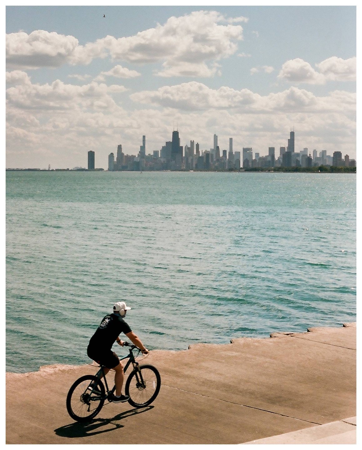 A person rides a bicycle along a concrete path next to a body of water with a city skyline in the distance.
