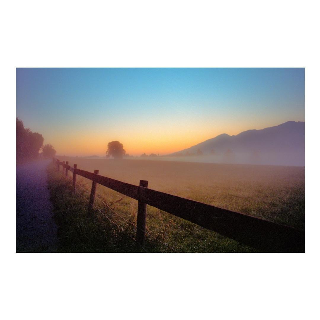 A wooden fence runs alongside a path and a field with mountains and trees in the distance under a colorful sky.