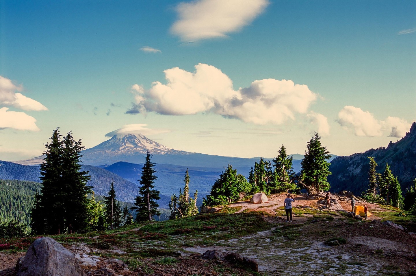 A person stands near a tent on a mountain with a snow-capped peak in the distance.