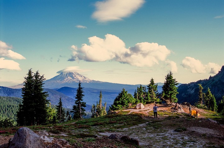 A person stands near a tent on a mountain with a snow-capped peak in the distance.