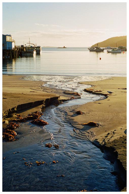 A small stream flows across a sandy beach towards a calm body of water with boats docked near a building and a distant hill.