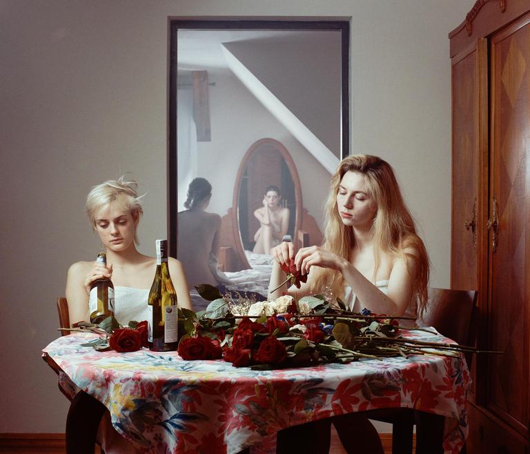 Two women sit at a table with roses and wine bottles, while a third woman is reflected in a mirror behind them.