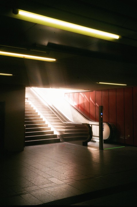 Sunlight streams down a long escalator and staircase in a subway station with red walls.