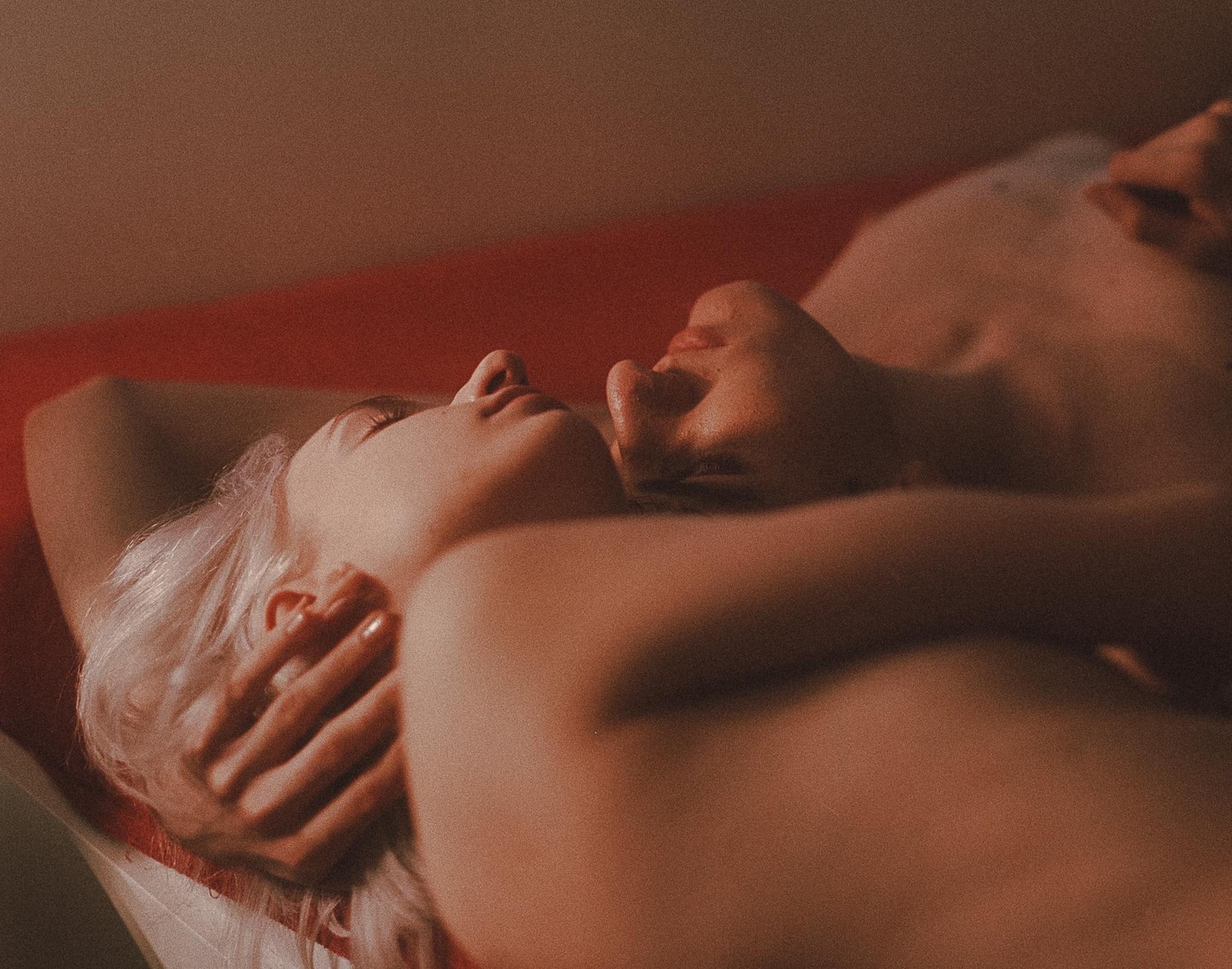 A couple lies intimately on a red surface, bathed in warm, soft light.