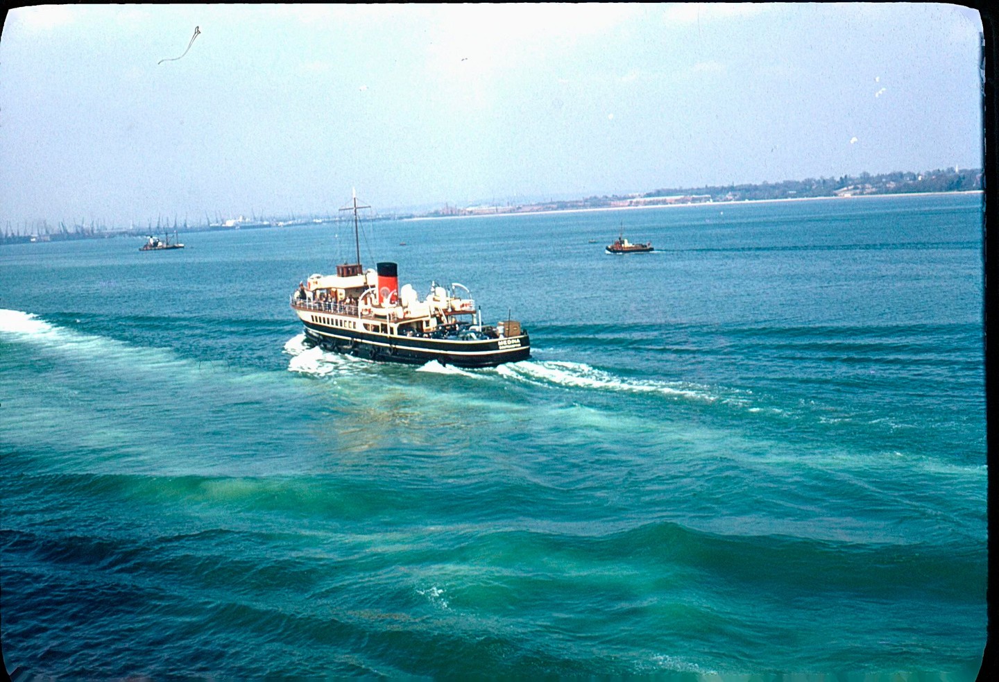 A ferry boat with passengers on deck moves through blue water, leaving a white wake behind it.