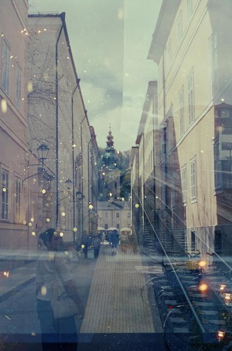 A street scene with buildings and people is overlaid with a reflection of a person and lights, creating a double exposure effect.