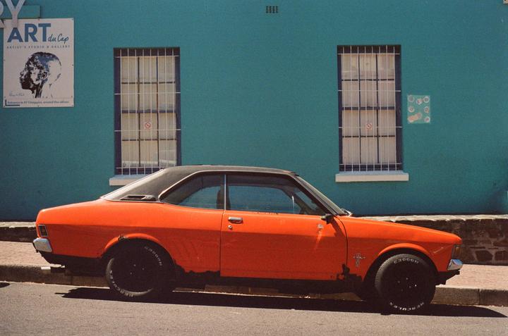 An orange car with a black roof is parked on a street next to a teal building with two windows.