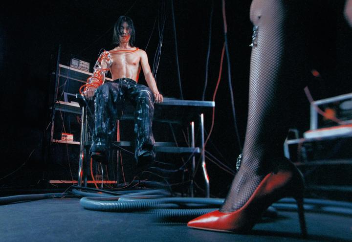 A man with wires attached to his arm sits on a metal structure while a woman's leg in a red high heel and fishnet stocking is in the foreground.