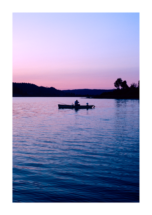 A man in a small boat fishes on a lake at sunset.