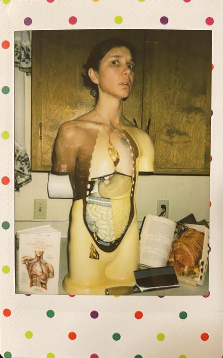 A woman stands next to an anatomical model with a book and a cooked ham on a counter.