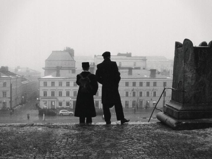 Two men stand on a cobblestone surface overlooking a city street with buildings in the distance on a hazy day.