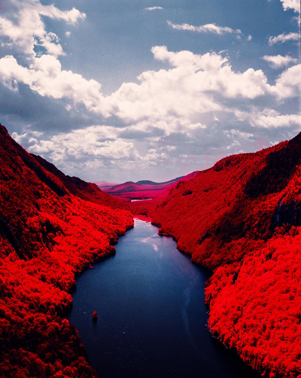 A river flows through a valley with red foliage on the mountainsides under a cloudy sky.