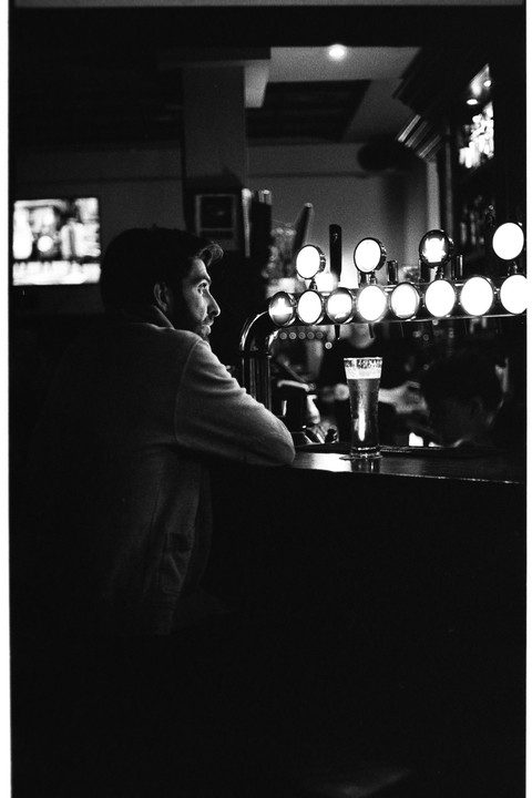 A man sits at a bar with a pint glass and beer taps in front of him.