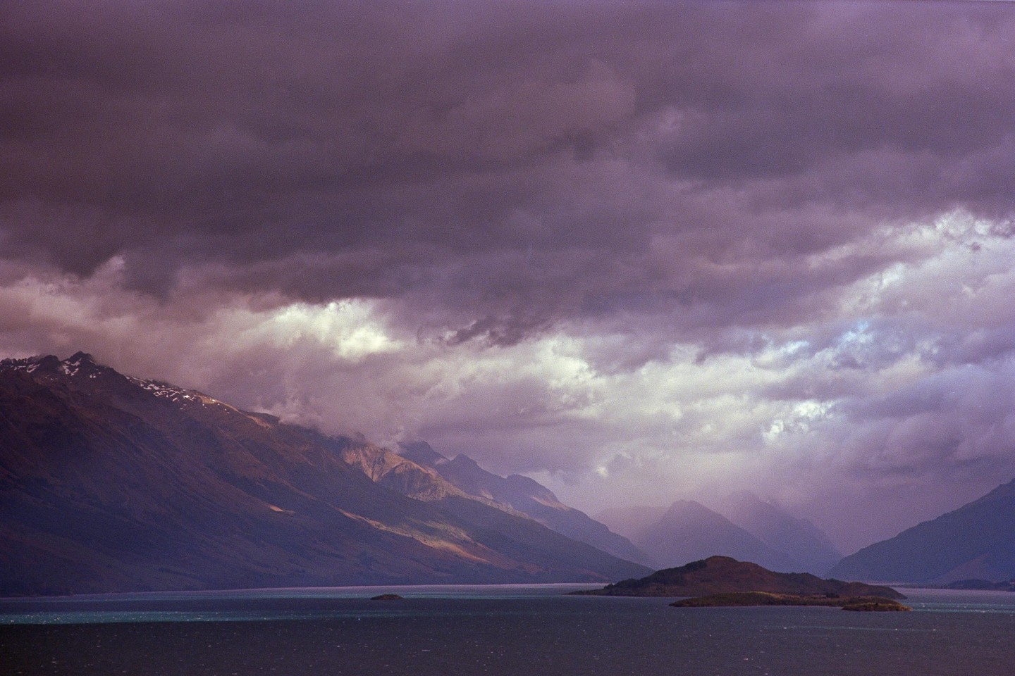 A dramatic landscape shows a lake with small islands and mountains under a cloudy sky.