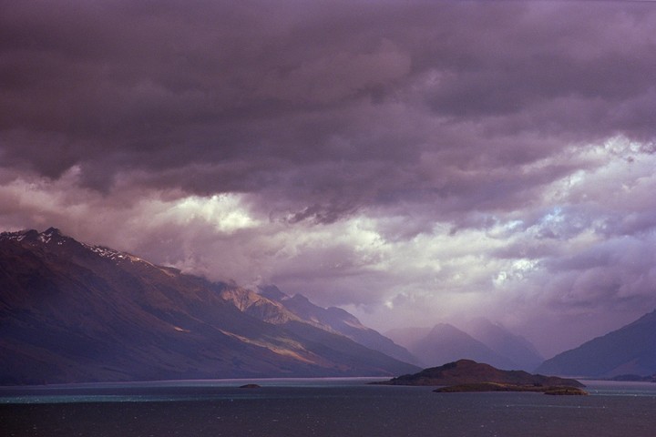 A dramatic landscape shows a lake with small islands and mountains under a cloudy sky.