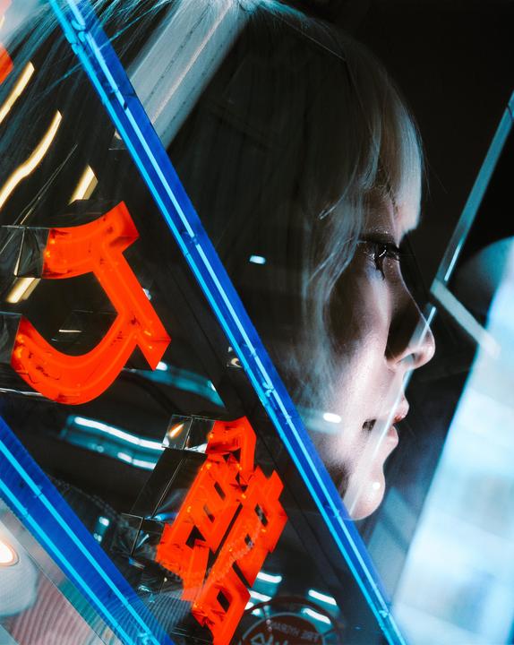 A woman's profile is visible through a window reflecting neon signs.