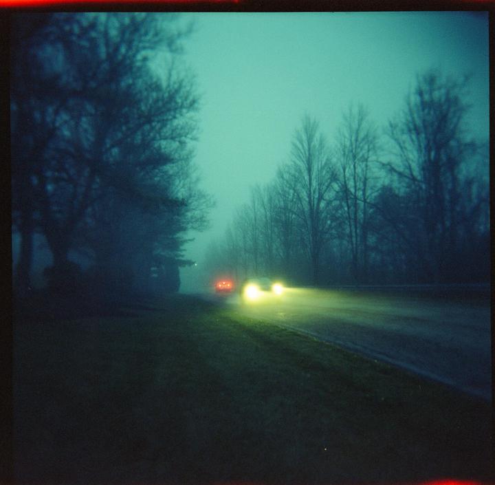 A car with bright headlights and red taillights drives down a road lined with trees in hazy conditions.