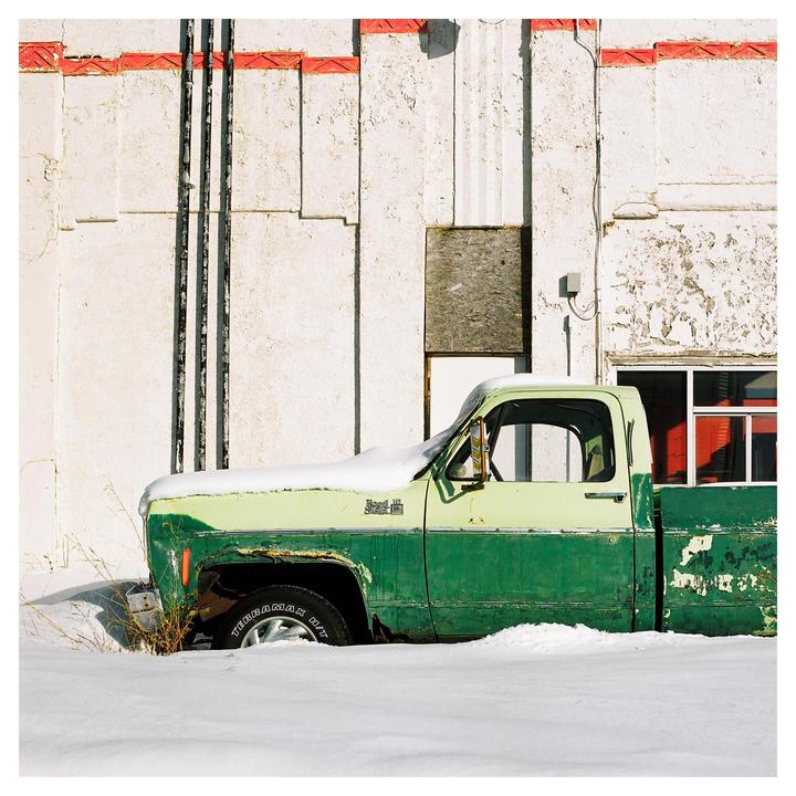 A snow-covered green and light green pickup truck is parked next to a white building with red trim.