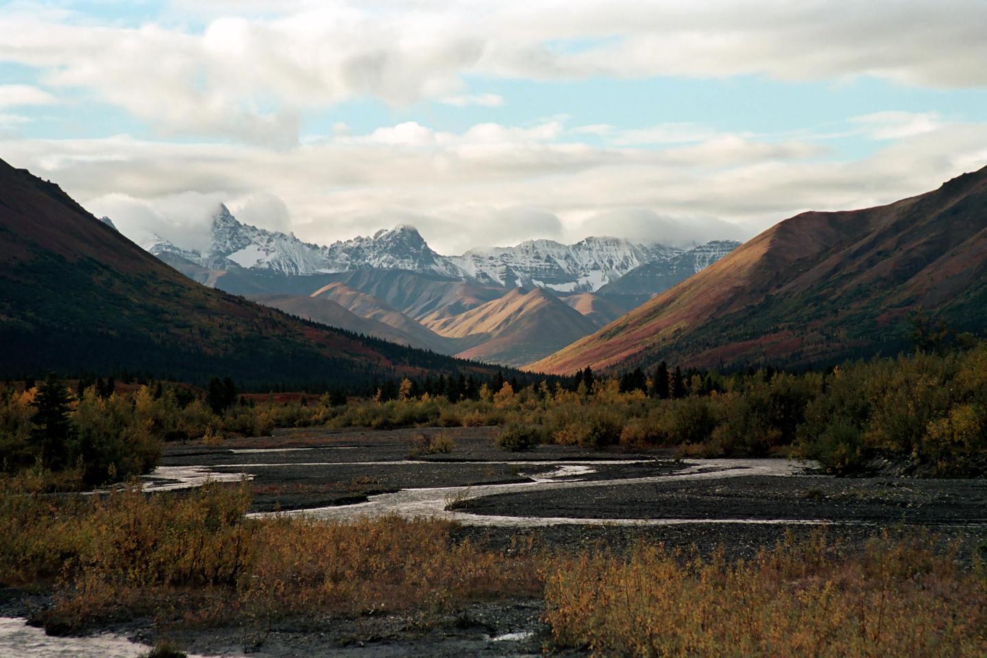 A river winds through a valley with autumn foliage and snow-capped mountains under a cloudy sky.