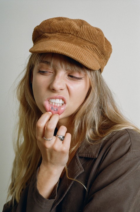 A woman wearing a corduroy hat pulls down her lower lip to reveal a "RUN" tattoo.