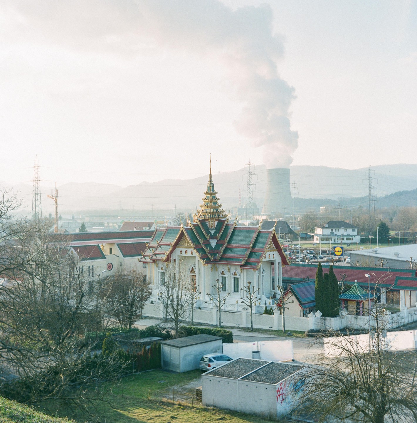 A temple with a tiered roof stands in a valley with a cooling tower emitting smoke in the background.