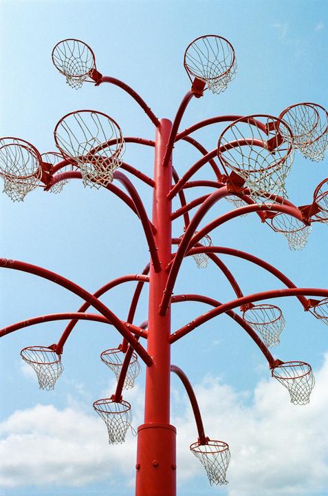 A red pole with multiple basketball hoops extends towards a blue sky with white clouds.