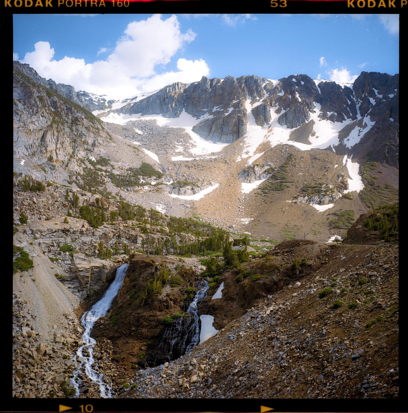 A waterfall cascades down a rocky mountain with snow-capped peaks under a blue sky.