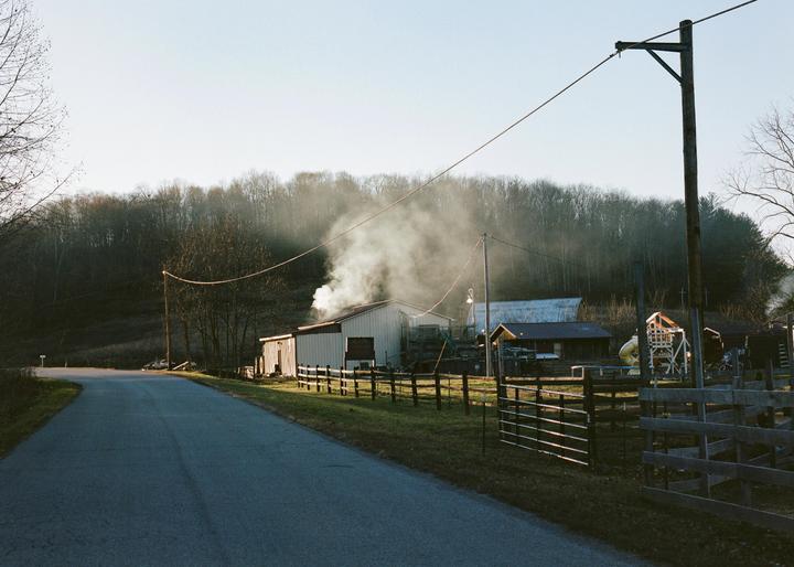 Smoke billows from a building on a farm next to a road with a forest in the background.